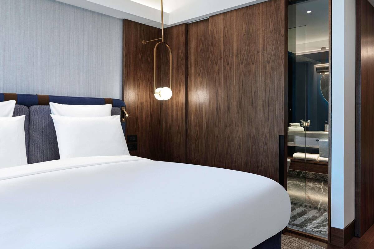 A modern hotel room with a large bed, wooden paneling, a glass shower enclosure, and a visible bathroom.