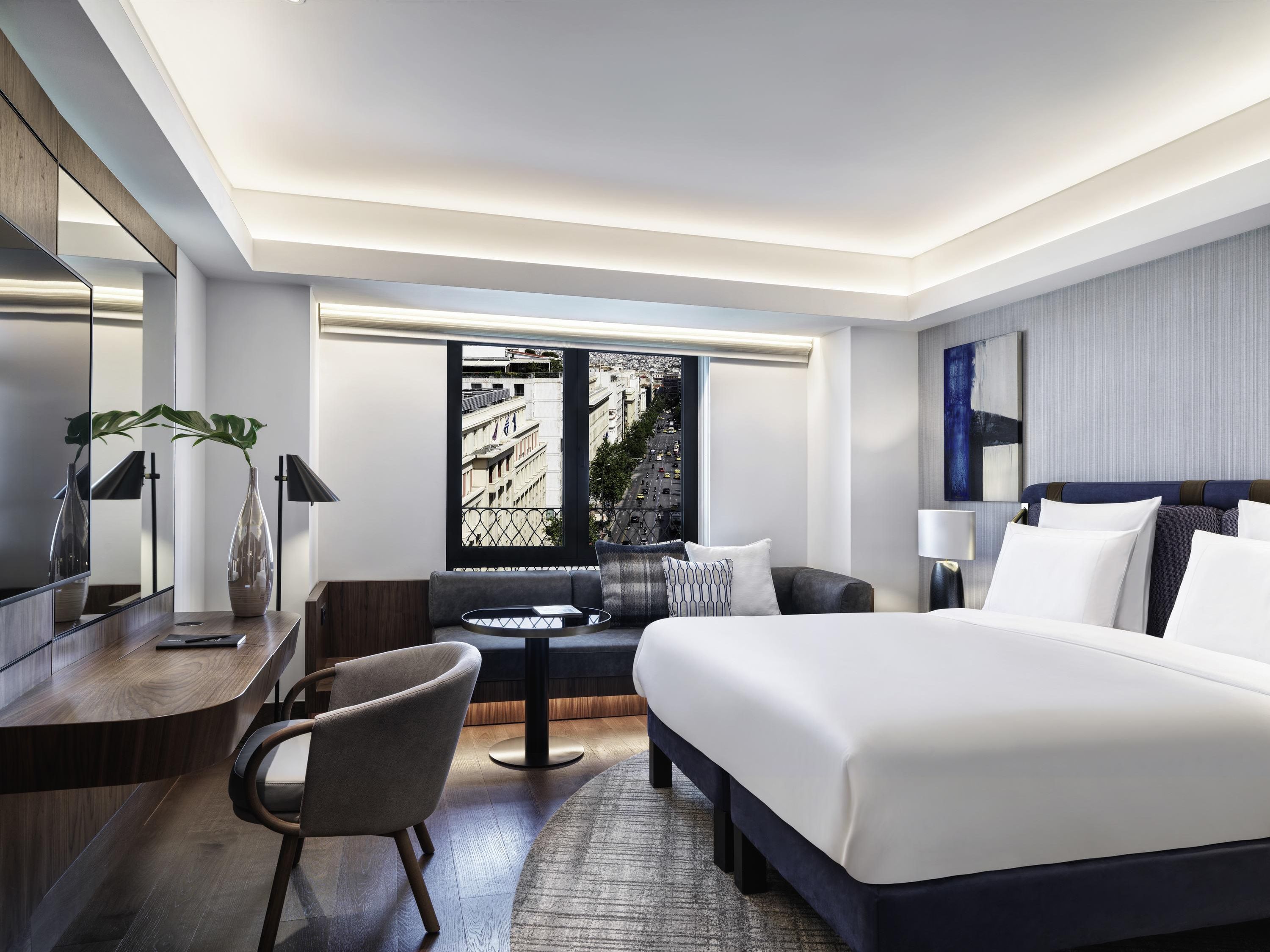 A modern hotel room with a large bed, a desk, a chair, a small table, and a window with a city view.