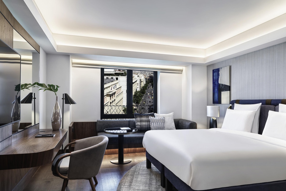 Una habitación de hotel moderna con una cama grande, un escritorio, una silla, una mesita y un ventanal con vistas a la ciudad.