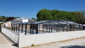 Een moderne kasconstructie met glazen dak, omgeven door een betonnen muur, en voorzien van zitplaatsen buiten.