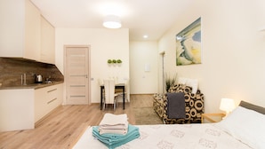 A compact hotel room with a kitchenette, a dining area, a bed with neatly folded towels, and a painting on the wall.