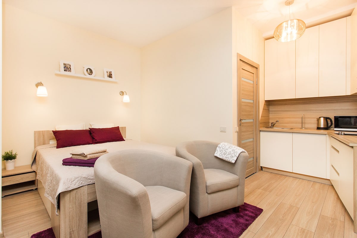 A bedroom with a bed, two armchairs, a small table, a nightstand, a purple rug, a wall-mounted shelf with photos, and a kitchenette with a microwave and kettle.