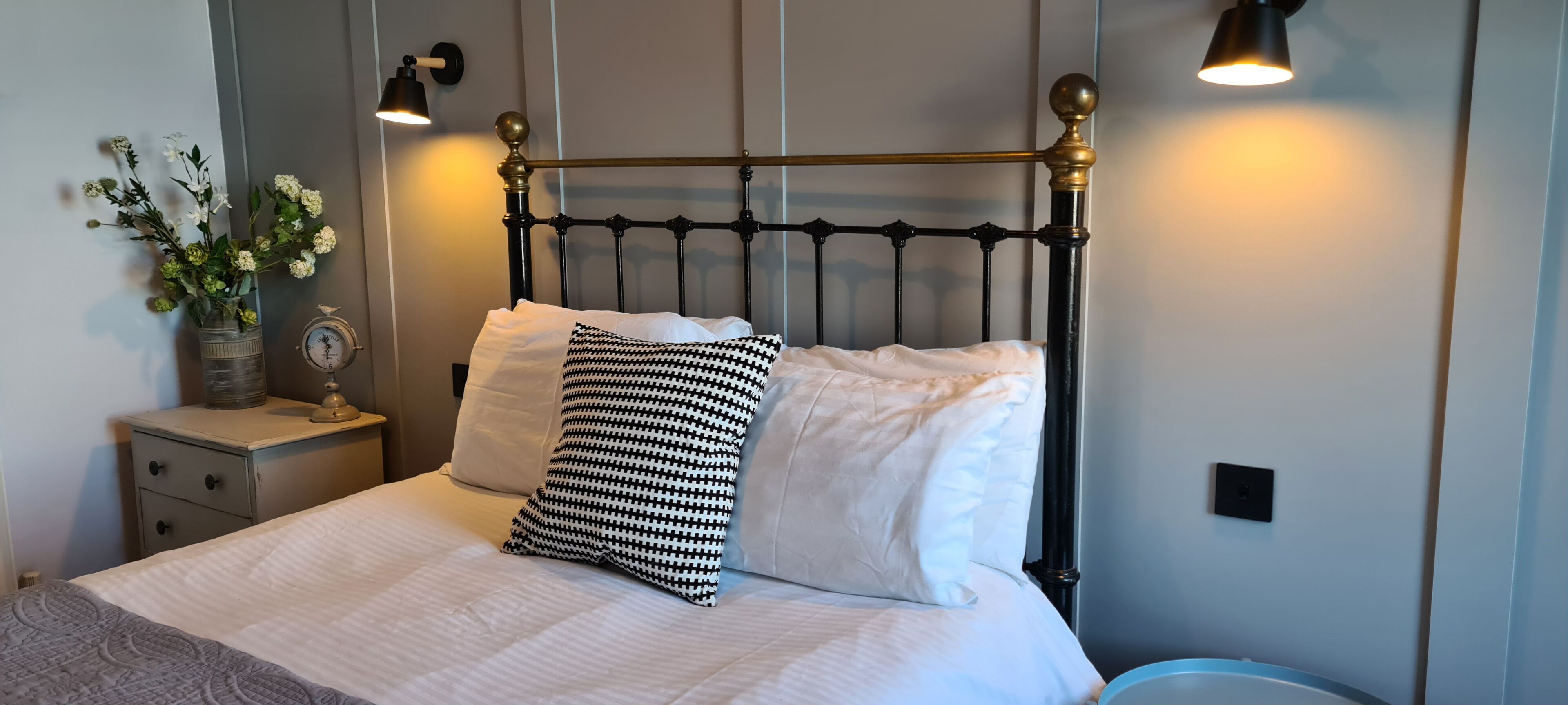 Een net opgemaakt bed met een zwart-wit gestreept kussen, een wit kussen en een witte deken. Er staat een nachtkastje met een klok en een bloevas op. De kamer heeft een modern design met een zwart metalen hoofdbord en twee wandlampen.
