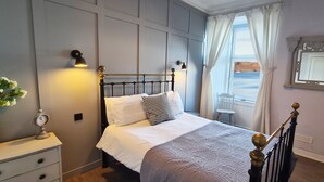 A bedroom with a bed, bedside lamps, a clock, and a view of the sea through a window. - 3 Oban - fantastic sea view apartment (Oban)