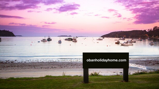 A serene waterfront scene with boats, a grassy foreground, and a sign with a website address. - 3 Oban - fantastic sea view apartment (Oban)