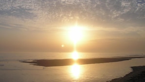 Une scène de plage où le soleil, bas à l’horizon, projette une lumière chaude sur l’eau et le sable.