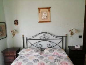Um quarto com estrutura de cama de metal, colcha estampada, um quadro na parede e uma pequena mesa de cabeceira com abajur.