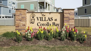 Une enseigne indiquant « The Villas at Corolla Bay », avec un pilier en pierre et des fleurs colorées devant.
