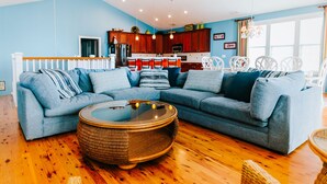A living room with a sectional sofa, a round wicker coffee table, and a kitchen in the background. -  6 bedroom-5.5 bathrooms in the heart of Garden City Beach and Heated Pool (Murrells Inlet)
