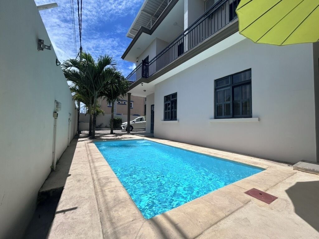 A swimming pool area with a clear blue pool, a concrete deck, and a white building with balconies.