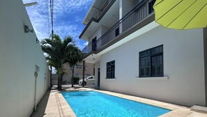 A swimming pool area with a clear blue pool, a concrete deck, and a white building with balconies.