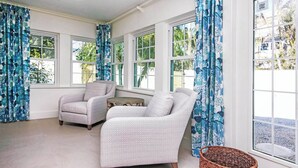Una habitación con dos sillones, una mesa redonda pequeña y una cesta de mimbre cerca de un ventanal con cortinas florales azules.