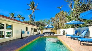 Una piscina con sillas de descanso y sombrillas, rodeada de palmeras y un edificio con grandes ventanales.