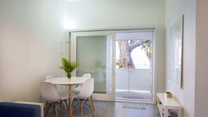 A modern living room with a dining area, a blue sofa, a white table with chairs, a potted plant, and a sliding glass door leading to a balcony with a tree view.