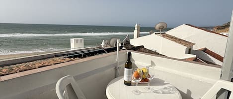 A terrace with a white table and chairs, a bottle of wine, and fruit. The terrace overlooks the ocean.