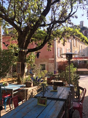 Un café en plein air avec des tables et des chaises en bois rustique, entouré de verdure et d’un grand arbre.