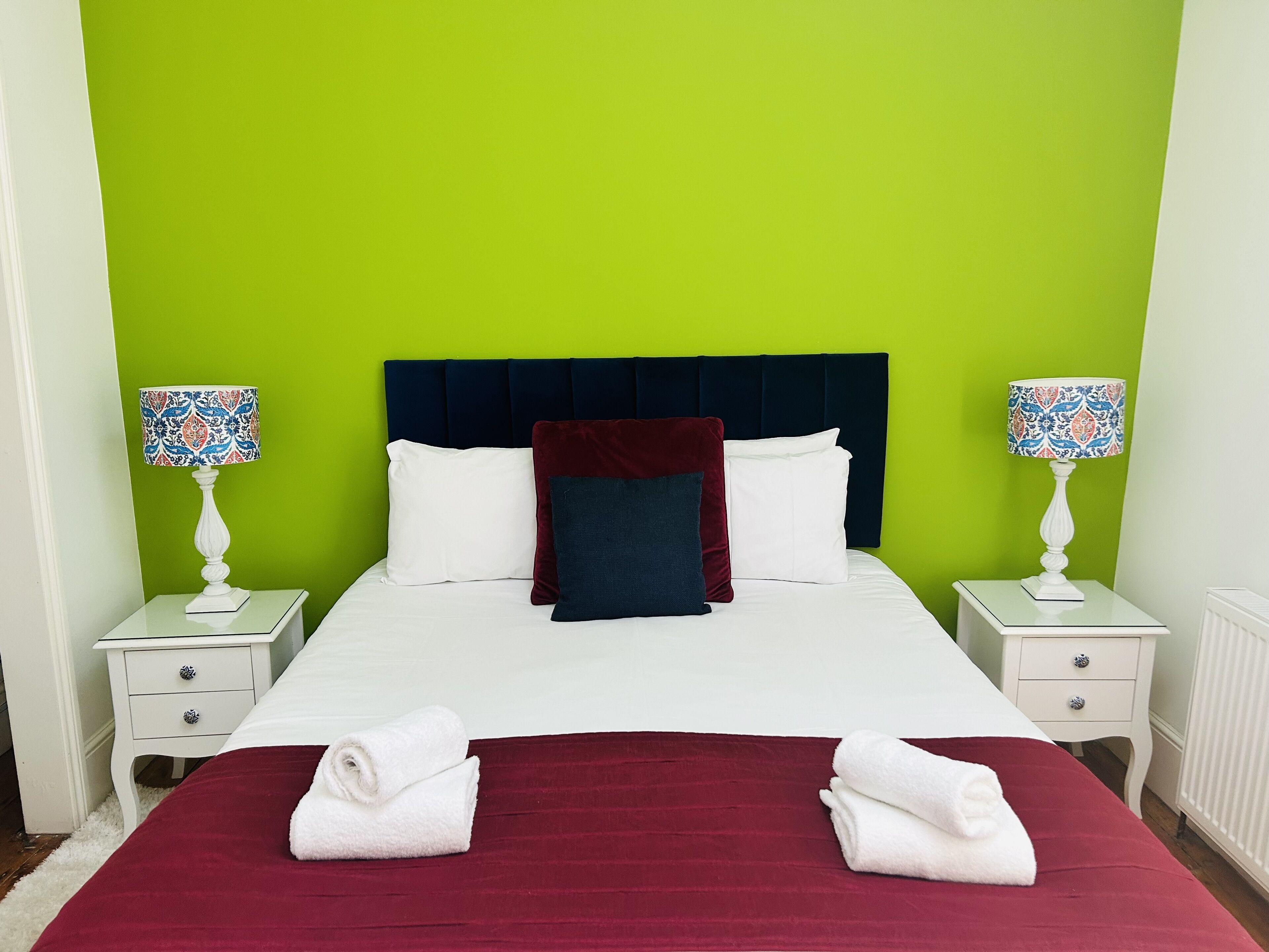 Un lit recouvert d’une couvre-lit blanche et rouge, deux tables de chevet blanches, deux lampes avec des abat-jour à motifs et un mur vert.