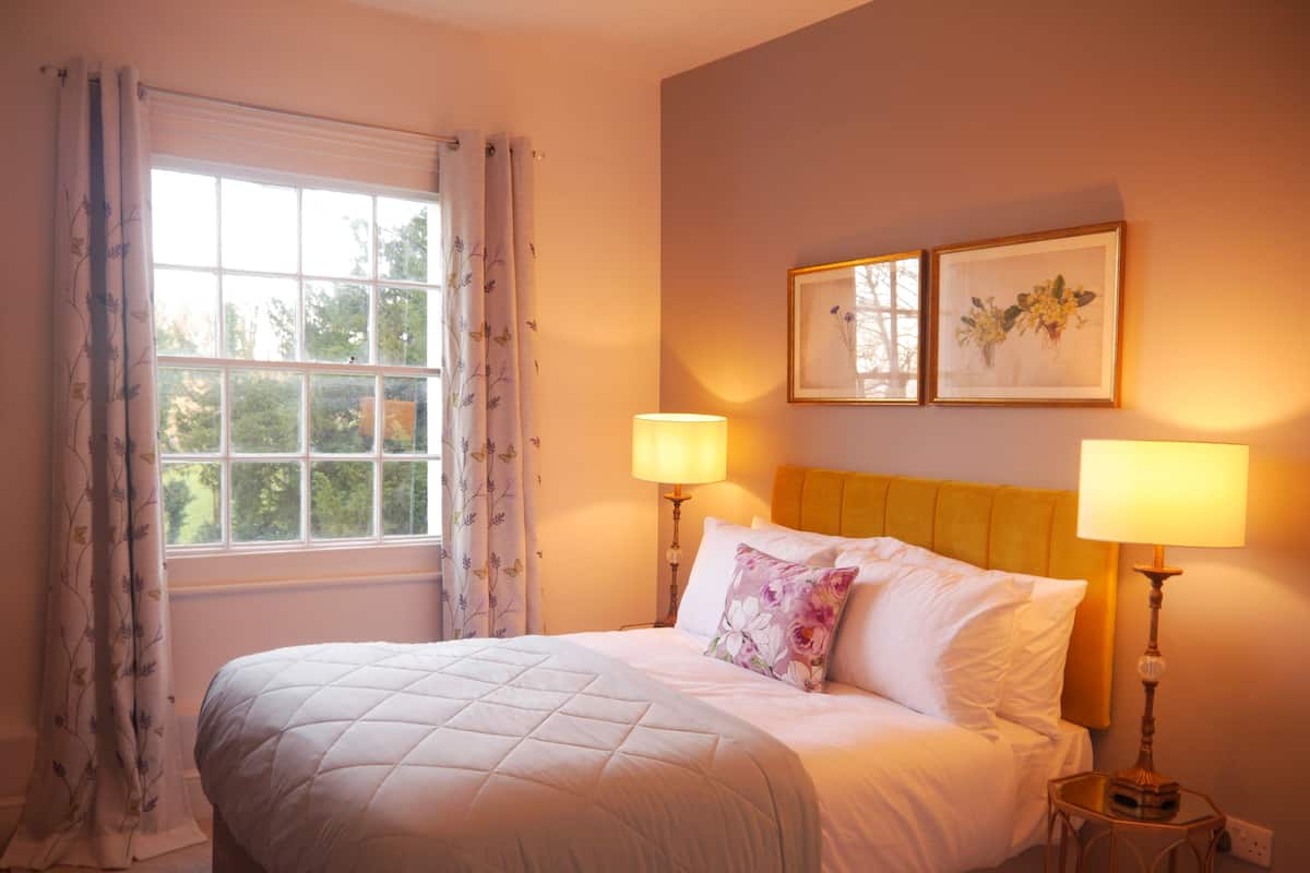 A neatly made bed with white linens and a floral pillow, two bedside lamps, and a window with curtains.
