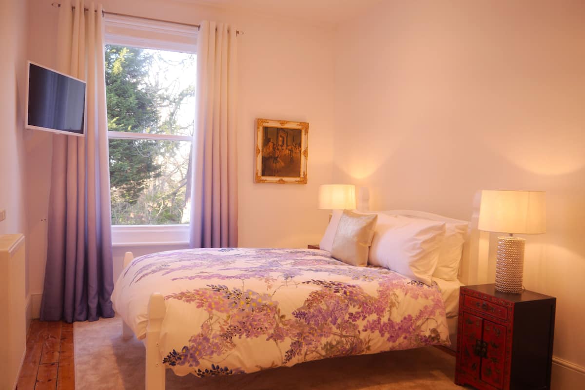 A bedroom with a bed, two bedside lamps, a TV mounted on the wall, and a window with curtains.