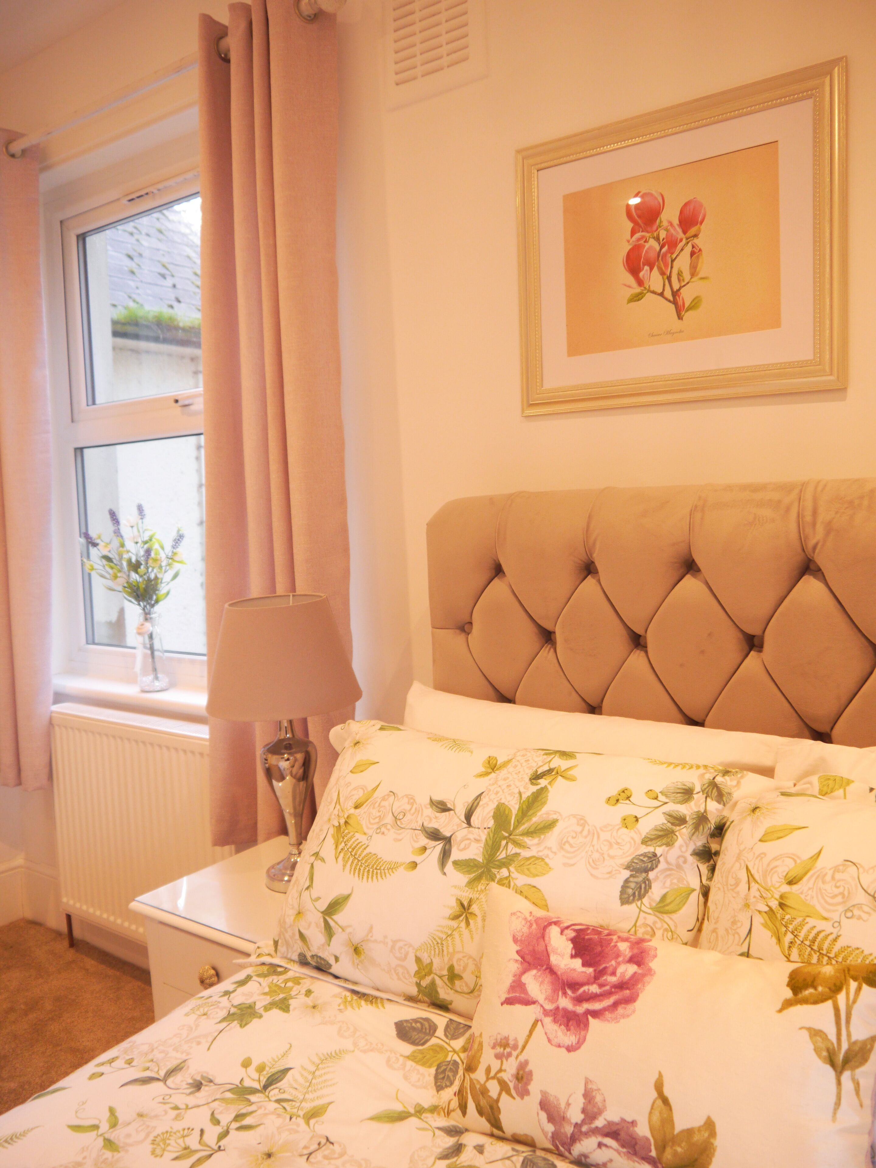 A bedroom with a tufted headboard, floral bedding, a bedside table with a lamp, and a window with curtains.