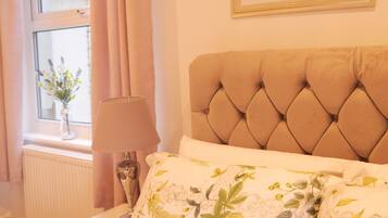 A bedroom with a tufted headboard, floral bedding, a bedside table with a lamp, and a window with curtains.