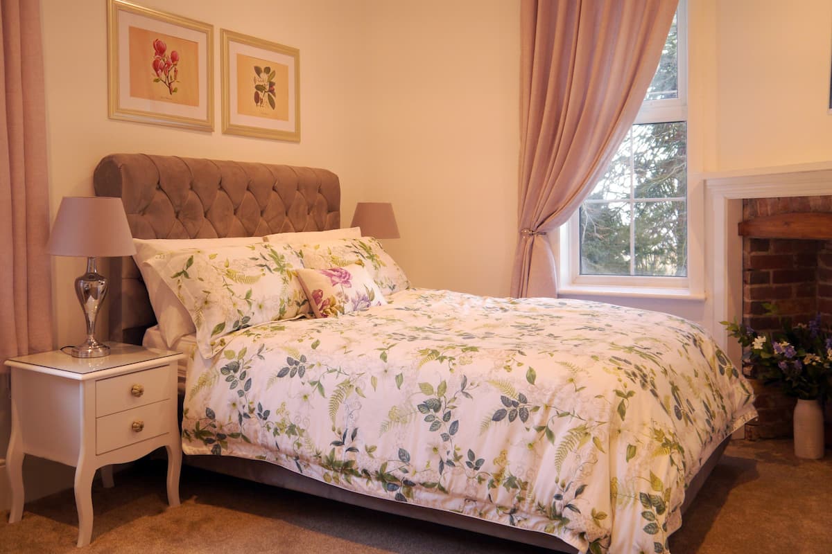 A bedroom with a tufted headboard, floral bedding, a bedside table with a lamp, and a window with curtains.