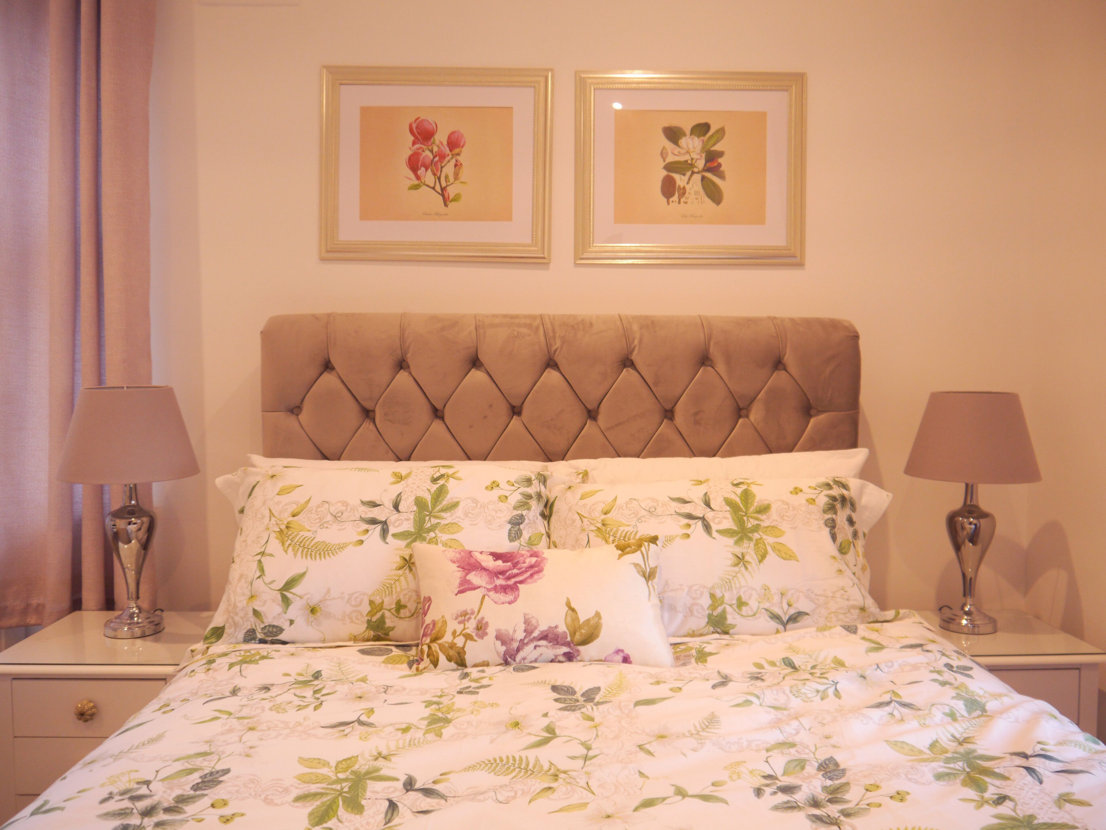 A bedroom with a tufted headboard, floral bedding, two bedside lamps, and two framed botanical prints on the wall.