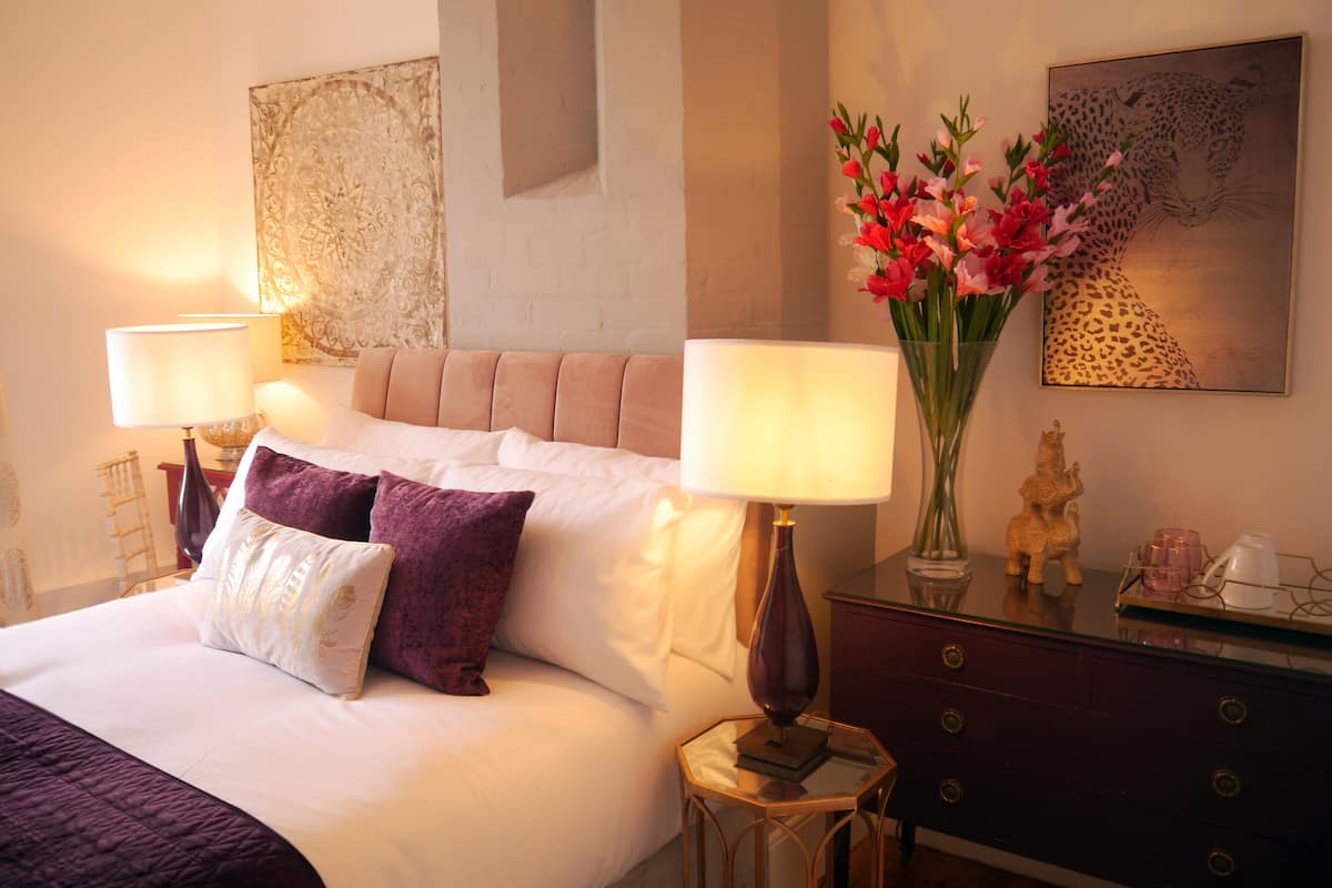 A neatly made bed with white and purple pillows, two bedside lamps, a vase with pink flowers, and a leopard print artwork on the wall.