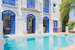 A poolside view of a white building with blue doors and windows, a covered outdoor seating area, and a palm tree. - The Pearl - Marbella B&B (Marbella)