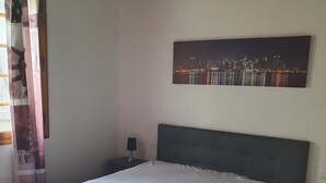 A bedroom with a large bed, a nightstand, a lamp, a cityscape painting, and a window with curtains. - Palavesa City (Porto-Vecchio)
