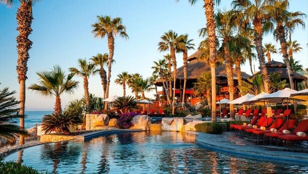 Zona de piscina en un resort con palmeras, sillas de descanso y un edificio de techo de paja.