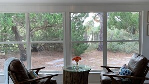 A room with two leather chairs, a glass table with a vase of flowers, and large windows offering a view of trees and a wooden deck.