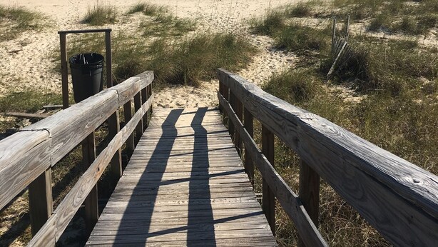 A wooden boardwalk leading to a beach with sand dunes and the ocean in the background.