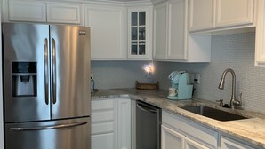 A modern kitchen with white cabinetry, a stainless steel refrigerator, a dishwasher, and a granite countertop.