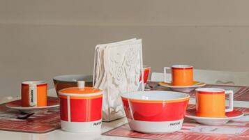 Una mesa dispuesta con tazas y cuencos naranja y blanco Pantone, un jarrón blanco decorativo, y un menú con diseño rojo y blanco.