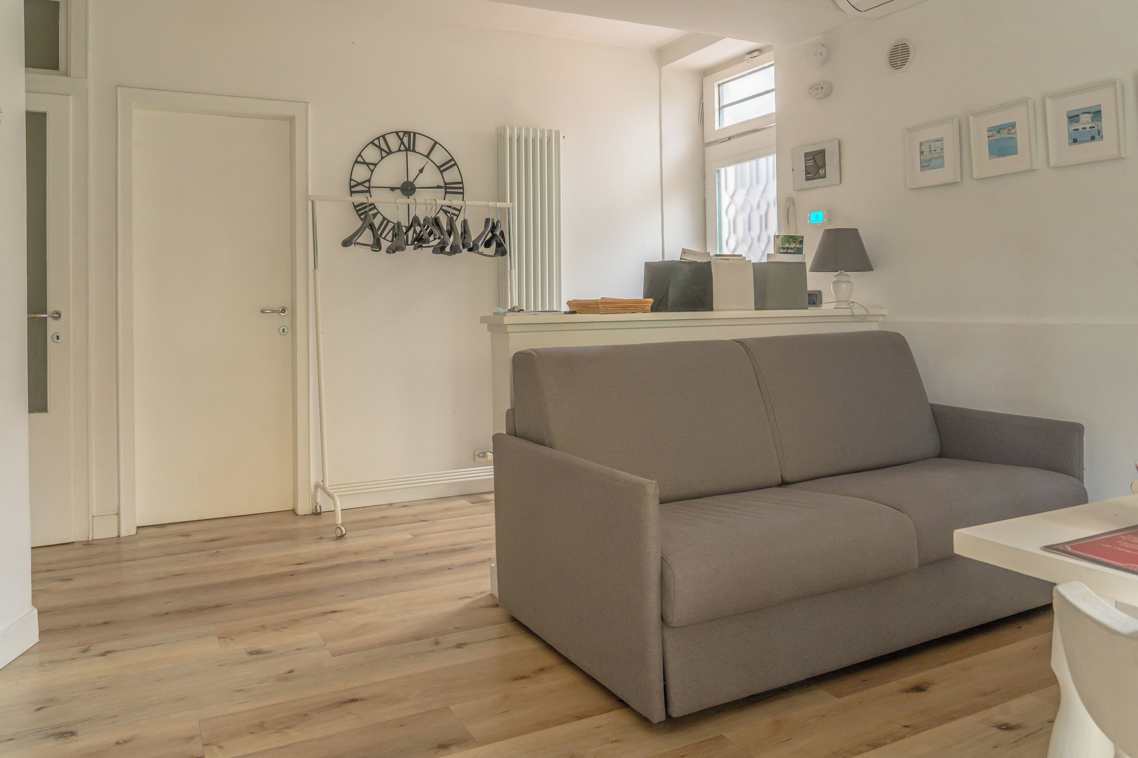 Un salon moderne avec un canapé gris, un plancher en bois, une horloge murale, un radiateur et une petite table avec une lampe.