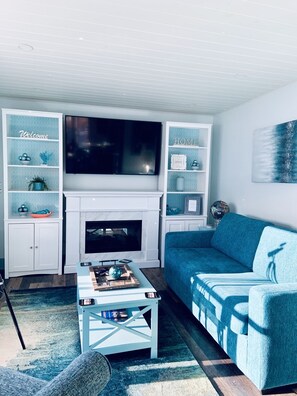 Un salon avec un téléviseur, une cheminée, un canapé bleu et des étagères ornées d’objets décoratifs.