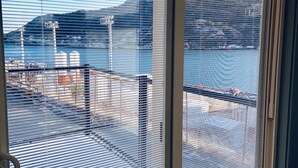 Une vue à travers une fenêtre munie de stores horizontaux, donnant sur une marina avec des bateaux et une chaîne de montagnes lointaine.