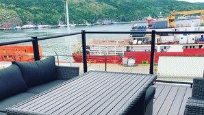 Une terrasse dotée d’une table et d’un espace salon, offrant une vue sur un port où sont amarrés des bateaux, dont un navire rouge.