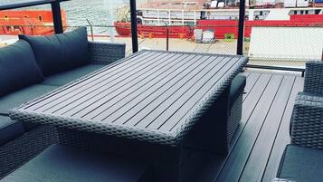 Une terrasse dotée d’une table et d’un espace salon, offrant une vue sur un port où sont amarrés des bateaux, dont un navire rouge.