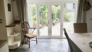 Una stanza con una porta vetrata scorrevole che si apre su un giardino, una sedia di legno, un tavolo bianco e un piccolo ripiano con un oggetto decorativo.