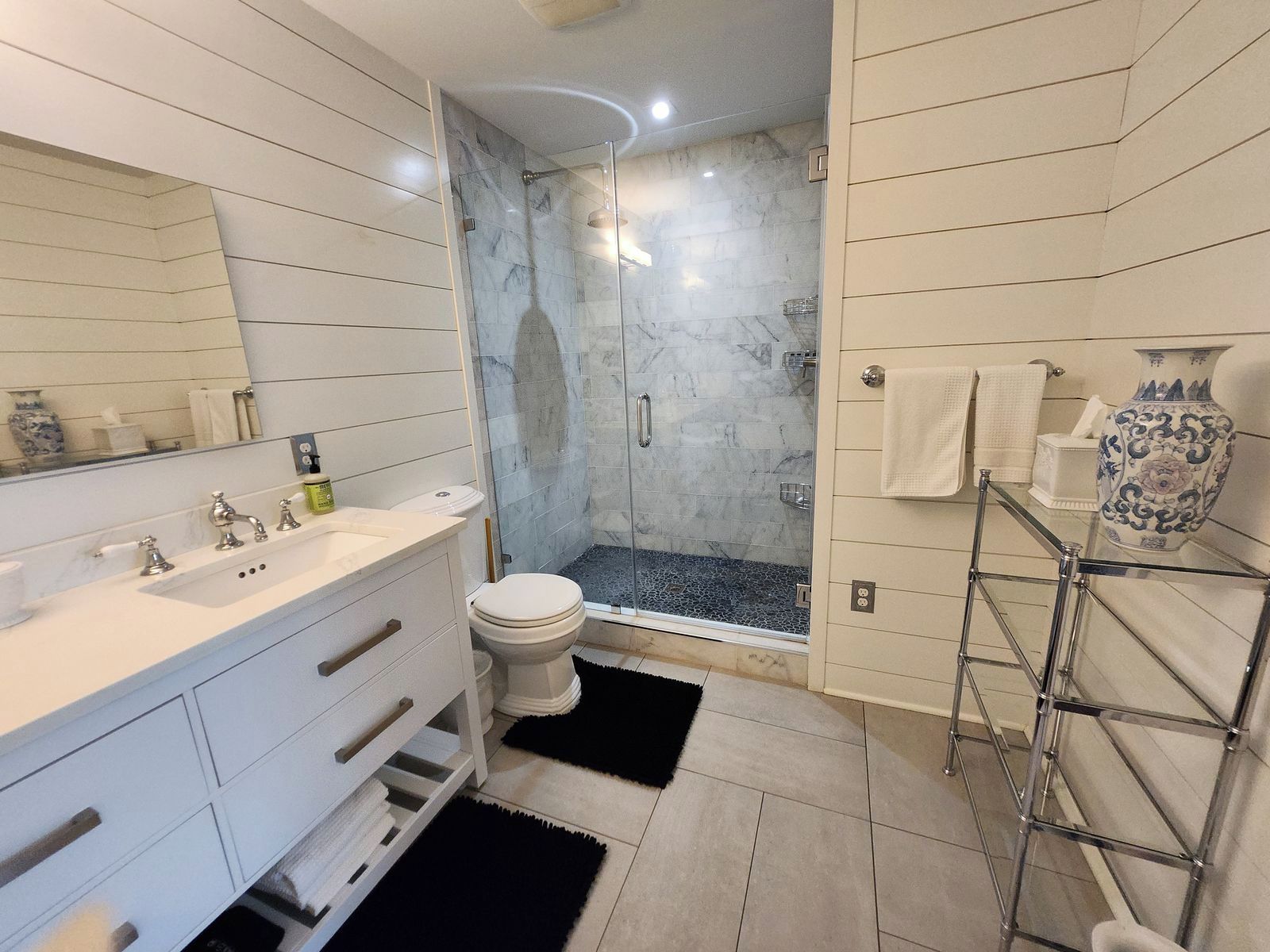 A modern bathroom with a glass-enclosed shower, a white vanity with a mirror, and a towel rack with a decorative vase.
