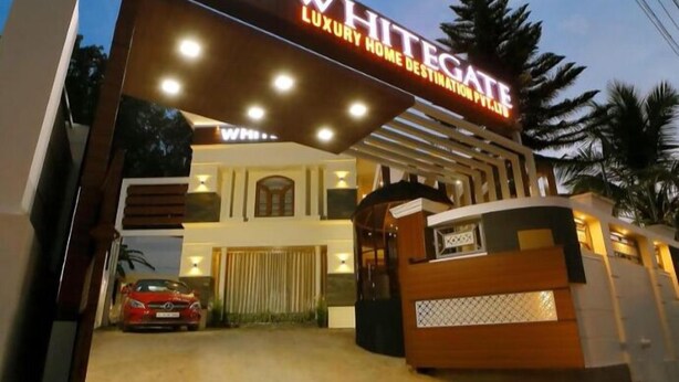 Une allée menant à une demeure de luxe, fermée par un portail, avec une voiture rouge garée à l’intérieur et un panneau bien éclairé.
