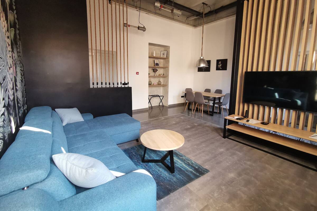 A modern living room with a blue sectional sofa, a round coffee table, a dining area with a wooden table and chairs, and a flat-screen TV mounted on a wooden panel.