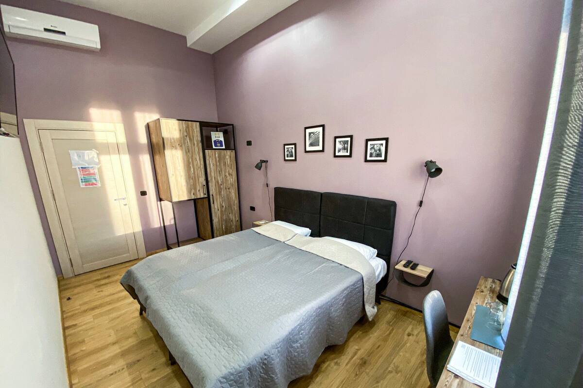 A bedroom with a bed, wooden wardrobe, bedside table, lamp, and a desk with a chair.