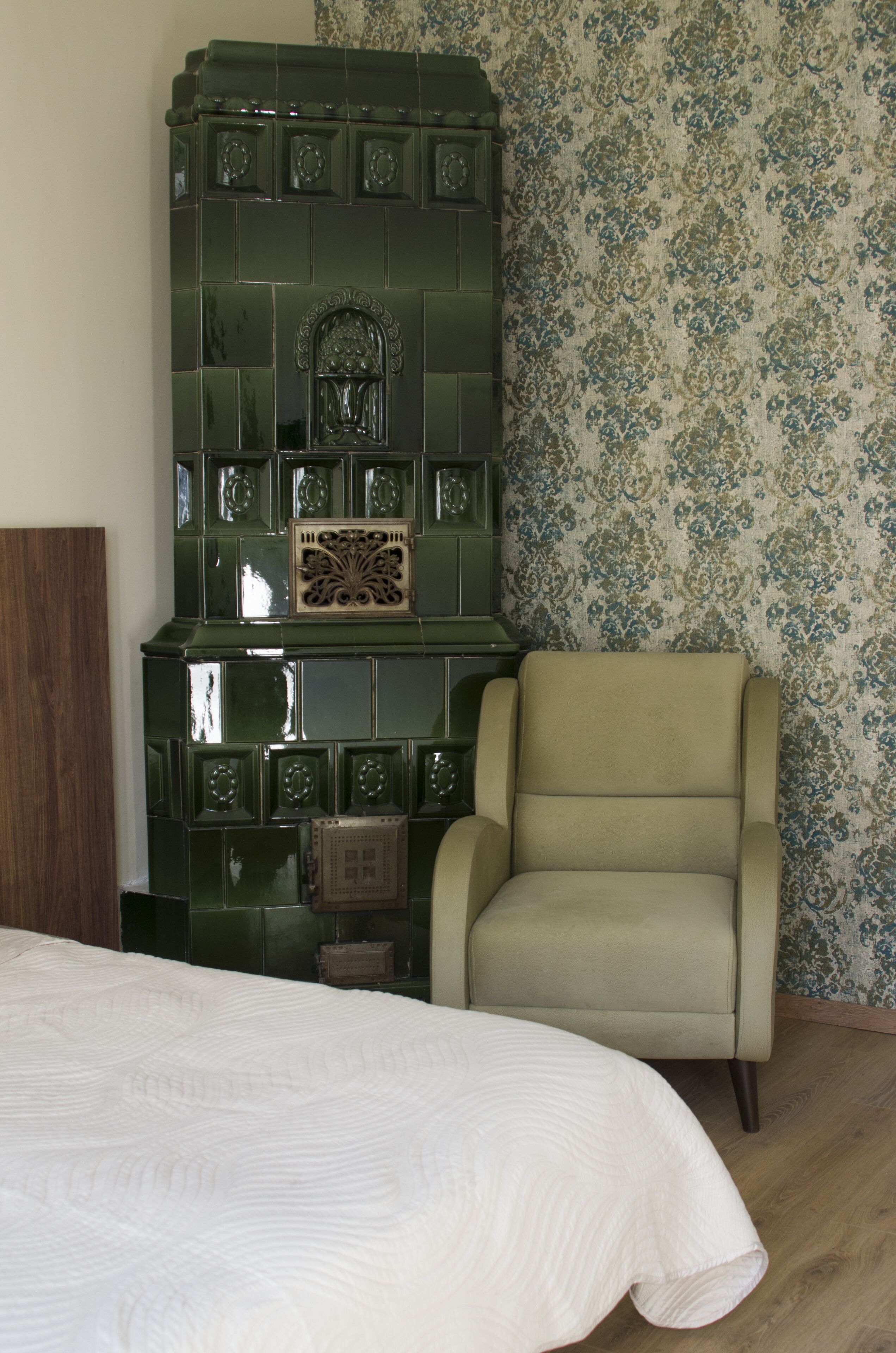 Een kamer met een groene betegelde open haard, een beige fauteuil en behang met een patroon.