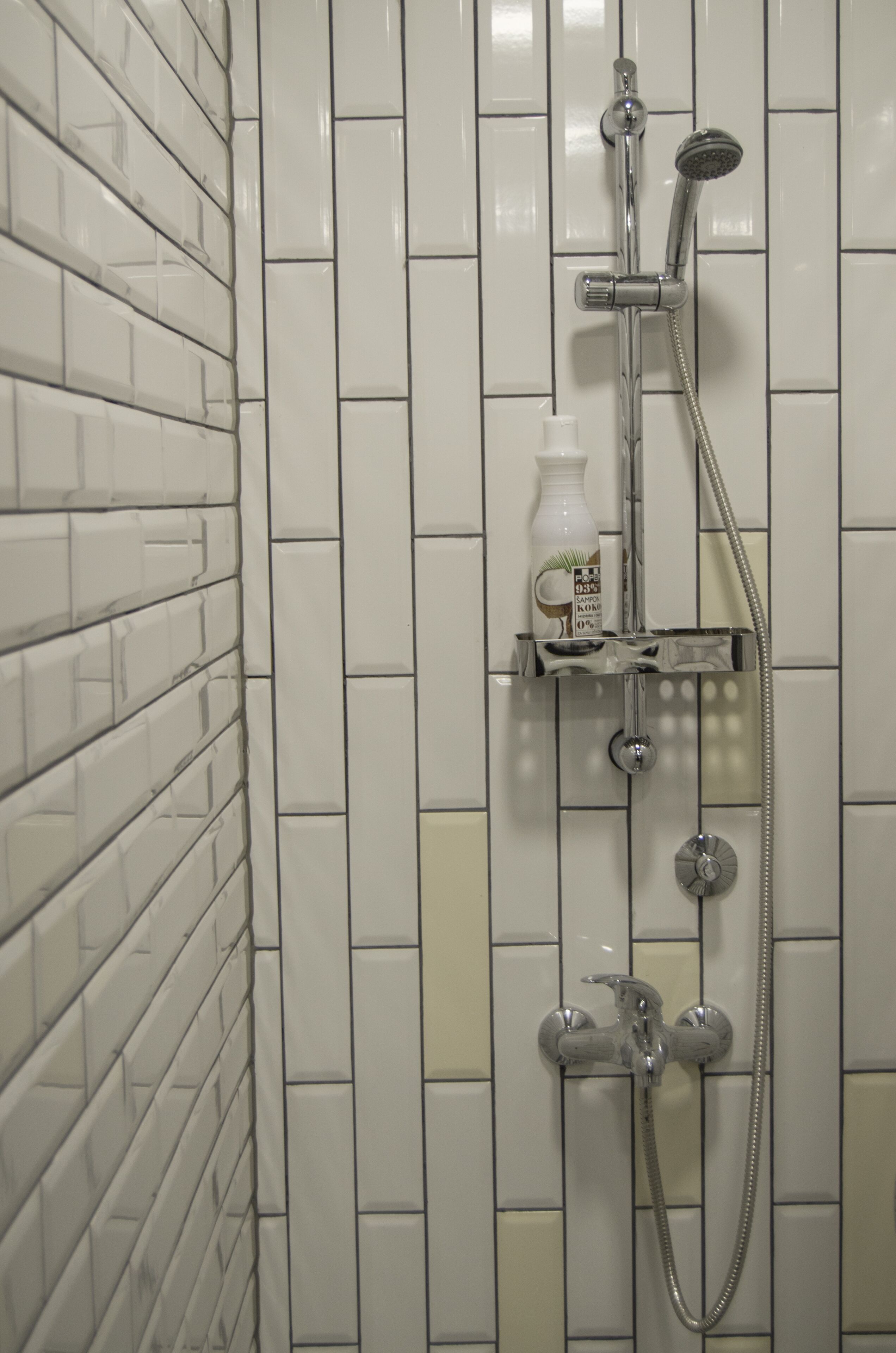 A bathroom with white tiled walls, a shower with a handheld showerhead, and a bottle of shampoo on a shelf.