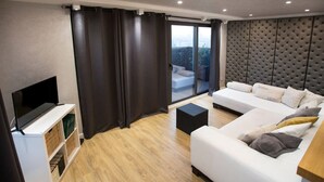A modern living room with a flat-screen TV, a white sofa set, a small black ottoman, and a balcony with a view.
