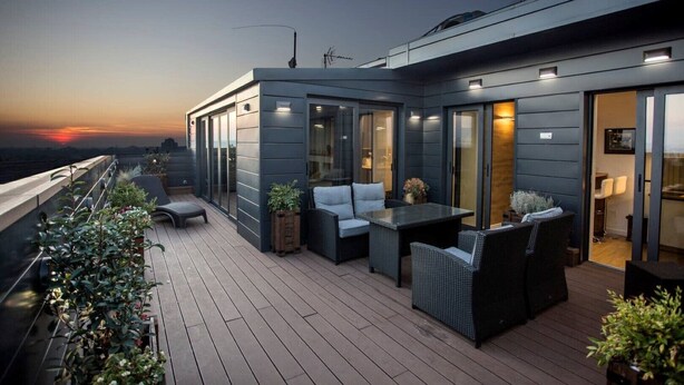A modern rooftop terrace with a wooden deck, outdoor seating, and a view of the sunset.