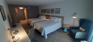 A hotel room with two beds, a desk, a chair, a lamp, and wall-mounted artwork.